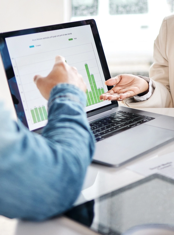 Two people discuss a bar graph displayed on a laptop screen, utilizing Business Intelligence for CFOs; one points at the chart while the other gestures with their hand.